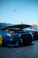A modified blue sports car with its hood open, showcasing the engine and custom features. The car is parked next to other vehicles on a concrete surface under a clear blue sky. Distinctive yellow rims and a lowered body kit are notable.