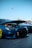 A modified blue sports car with its hood open, showcasing the engine and custom features. The car is parked next to other vehicles on a concrete surface under a clear blue sky. Distinctive yellow rims and a lowered body kit are notable.