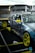 A modified sports car with a camo paint job and bright yellow accents, including rims and side mirrors, is positioned inside a tented area or garage. The car is equipped with performance parts such as a large rear spoiler, roll cage, and racing decals. The surroundings include workshop equipment and tires.