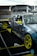 A modified sports car with a camo paint job and bright yellow accents, including rims and side mirrors, is positioned inside a tented area or garage. The car is equipped with performance parts such as a large rear spoiler, roll cage, and racing decals. The surroundings include workshop equipment and tires.