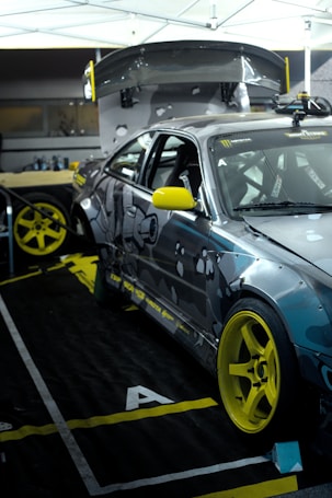 A modified sports car with a camo paint job and bright yellow accents, including rims and side mirrors, is positioned inside a tented area or garage. The car is equipped with performance parts such as a large rear spoiler, roll cage, and racing decals. The surroundings include workshop equipment and tires.