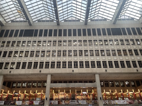 A large interior space featuring a grid-patterned, skylit ceiling with a modern architectural design. The facade is composed of multiple rows of square windows and geometric patterns. Below, there are illuminated signs and food stalls, suggesting a shopping or dining area.