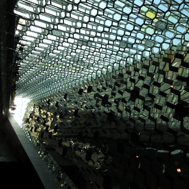 A complex pattern of hexagonal glass panels forms the structure of a ceiling or wall, creating an intricate play of light and shadow. The panels reflect various shades of green and blue, adding depth and a sense of architectural modernism.