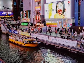 A bustling urban scene with a vibrant atmosphere, featuring a crowded riverside area with people walking along a promenade. A bright yellow tour boat with passengers is on the river. Large, colorful advertisements and billboards light up the surrounding buildings, reflecting off the water. The area is lively, with various shops and signs in both Japanese and English.