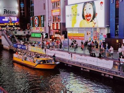 A bustling urban scene with a vibrant atmosphere, featuring a crowded riverside area with people walking along a promenade. A bright yellow tour boat with passengers is on the river. Large, colorful advertisements and billboards light up the surrounding buildings, reflecting off the water. The area is lively, with various shops and signs in both Japanese and English.