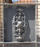 An intricate metal design is set within a rectangular frame. The design features ornate floral and scroll patterns, with a weathered and slightly rusted texture. The background appears to be a grid or mesh, and sunlight casts shadows on the surface.