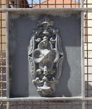 An intricate metal design is set within a rectangular frame. The design features ornate floral and scroll patterns, with a weathered and slightly rusted texture. The background appears to be a grid or mesh, and sunlight casts shadows on the surface.