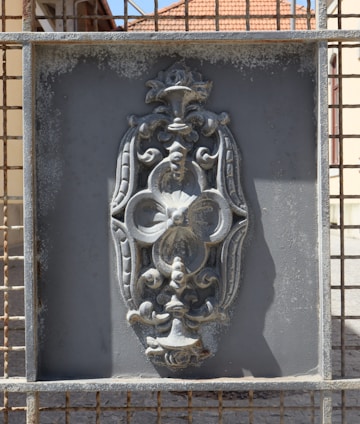 An intricate metal design is set within a rectangular frame. The design features ornate floral and scroll patterns, with a weathered and slightly rusted texture. The background appears to be a grid or mesh, and sunlight casts shadows on the surface.