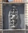 An intricate metal design is set within a rectangular frame. The design features ornate floral and scroll patterns, with a weathered and slightly rusted texture. The background appears to be a grid or mesh, and sunlight casts shadows on the surface.