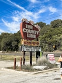 A large vintage-style red sign shaped like a brisket advertises a barbecue restaurant called 'The County Line.' The sign features pigs on top and a message encouraging patrons to 'eat more barbeque here or at home.' A smaller sign nearby announces 'Happy Hour' details. Surrounding the signs are patches of grass and trees under a clear blue sky with wispy clouds. A person with a camera is partially visible in the foreground.