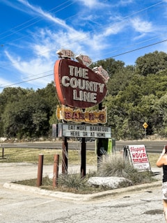A large vintage-style red sign shaped like a brisket advertises a barbecue restaurant called 'The County Line.' The sign features pigs on top and a message encouraging patrons to 'eat more barbeque here or at home.' A smaller sign nearby announces 'Happy Hour' details. Surrounding the signs are patches of grass and trees under a clear blue sky with wispy clouds. A person with a camera is partially visible in the foreground.