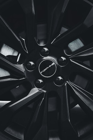 A close-up view of a car wheel rim with a sleek, modern design featuring sharp lines and a glossy black finish. The center cap includes the branding with a circular arrangement of lug nuts.