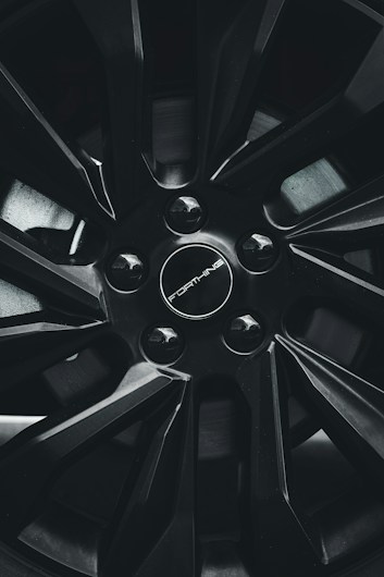 A close-up view of a car wheel rim with a sleek, modern design featuring sharp lines and a glossy black finish. The center cap includes the branding with a circular arrangement of lug nuts.