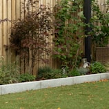 A garden scene featuring a wooden fence in the background, surrounded by various green plants and a tree with reddish-brown leaves. There is a small patch of lawn in the foreground, edged by a light gray stone border. On the right side, a wicker basket and a reflective, metallic garden ornament are visible.