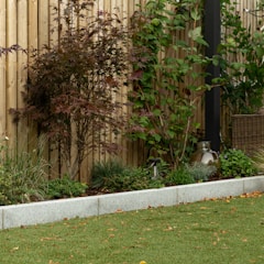 A garden scene featuring a wooden fence in the background, surrounded by various green plants and a tree with reddish-brown leaves. There is a small patch of lawn in the foreground, edged by a light gray stone border. On the right side, a wicker basket and a reflective, metallic garden ornament are visible.