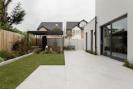 A modern backyard featuring a spacious, tiled patio area adjacent to a large rectangular patch of well-maintained grass. A pergola with outdoor seating sits at the end of the garden, surrounded by plants and trees that provide a touch of greenery. The surrounding fence is wooden, separating the space from neighboring homes. The main building includes large glass doors that open onto the patio.