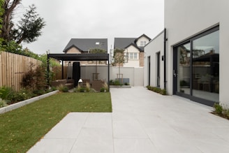 A modern backyard featuring a spacious, tiled patio area adjacent to a large rectangular patch of well-maintained grass. A pergola with outdoor seating sits at the end of the garden, surrounded by plants and trees that provide a touch of greenery. The surrounding fence is wooden, separating the space from neighboring homes. The main building includes large glass doors that open onto the patio.