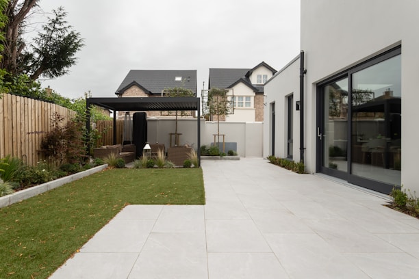 A modern backyard featuring a spacious, tiled patio area adjacent to a large rectangular patch of well-maintained grass. A pergola with outdoor seating sits at the end of the garden, surrounded by plants and trees that provide a touch of greenery. The surrounding fence is wooden, separating the space from neighboring homes. The main building includes large glass doors that open onto the patio.