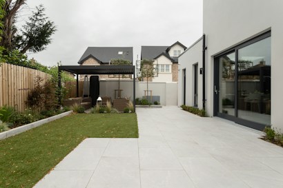 A modern backyard featuring a spacious, tiled patio area adjacent to a large rectangular patch of well-maintained grass. A pergola with outdoor seating sits at the end of the garden, surrounded by plants and trees that provide a touch of greenery. The surrounding fence is wooden, separating the space from neighboring homes. The main building includes large glass doors that open onto the patio.