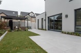 A modern outdoor patio area with a well-maintained lawn and a sleek white building. The scene includes a pergola with seating, surrounded by small shrubs. In the background, multi-story houses with modern architecture are visible.