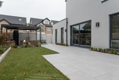 A modern outdoor patio area with a well-maintained lawn and a sleek white building. The scene includes a pergola with seating, surrounded by small shrubs. In the background, multi-story houses with modern architecture are visible.