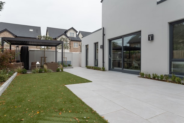 A modern outdoor patio area with a well-maintained lawn and a sleek white building. The scene includes a pergola with seating, surrounded by small shrubs. In the background, multi-story houses with modern architecture are visible.