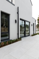 A modern, minimalist exterior view of a building with large glass windows and clean, white walls. The ground is paved with large light-colored tiles, and there is a row of small shrubs planted along the building's edge. A black drainpipe is attached vertically to the wall.