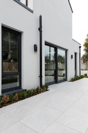 A modern, minimalist exterior view of a building with large glass windows and clean, white walls. The ground is paved with large light-colored tiles, and there is a row of small shrubs planted along the building's edge. A black drainpipe is attached vertically to the wall.
