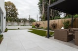 An outdoor patio features a spacious tiled area leading to a section with artificial grass. Wicker furniture, including a sofa and chairs, is positioned beneath a modern pergola. A wooden fence encloses the yard, while trees and bushes add greenery to the setting. The sky appears overcast.