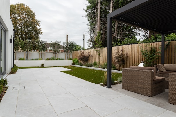 An outdoor patio features a spacious tiled area leading to a section with artificial grass. Wicker furniture, including a sofa and chairs, is positioned beneath a modern pergola. A wooden fence encloses the yard, while trees and bushes add greenery to the setting. The sky appears overcast.