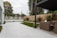 An outdoor patio features a spacious tiled area leading to a section with artificial grass. Wicker furniture, including a sofa and chairs, is positioned beneath a modern pergola. A wooden fence encloses the yard, while trees and bushes add greenery to the setting. The sky appears overcast.