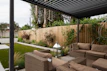 A modern outdoor patio area features wicker furniture with beige cushions arranged under a pergola. The setting includes a well-maintained garden with green grass, shrubs, and a wooden fence adorned with climbing plants. A metallic lantern stands nearby, adding an element of decor.