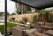 A modern outdoor patio area features wicker furniture with beige cushions arranged under a pergola. The setting includes a well-maintained garden with green grass, shrubs, and a wooden fence adorned with climbing plants. A metallic lantern stands nearby, adding an element of decor.