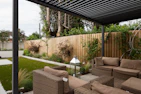 A modern outdoor patio area features wicker furniture with beige cushions arranged under a pergola. The setting includes a well-maintained garden with green grass, shrubs, and a wooden fence adorned with climbing plants. A metallic lantern stands nearby, adding an element of decor.