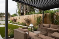 A modern outdoor patio area features wicker furniture with beige cushions arranged under a pergola. The setting includes a well-maintained garden with green grass, shrubs, and a wooden fence adorned with climbing plants. A metallic lantern stands nearby, adding an element of decor.