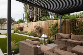 A modern outdoor patio area features wicker furniture with beige cushions arranged under a pergola. The setting includes a well-maintained garden with green grass, shrubs, and a wooden fence adorned with climbing plants. A metallic lantern stands nearby, adding an element of decor.
