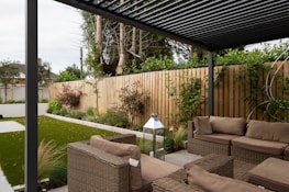 A modern outdoor patio area features wicker furniture with beige cushions arranged under a pergola. The setting includes a well-maintained garden with green grass, shrubs, and a wooden fence adorned with climbing plants. A metallic lantern stands nearby, adding an element of decor.