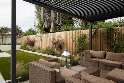 A modern outdoor patio area features wicker furniture with beige cushions arranged under a pergola. The setting includes a well-maintained garden with green grass, shrubs, and a wooden fence adorned with climbing plants. A metallic lantern stands nearby, adding an element of decor.