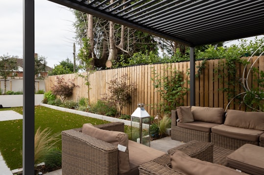 A modern outdoor patio area features wicker furniture with beige cushions arranged under a pergola. The setting includes a well-maintained garden with green grass, shrubs, and a wooden fence adorned with climbing plants. A metallic lantern stands nearby, adding an element of decor.
