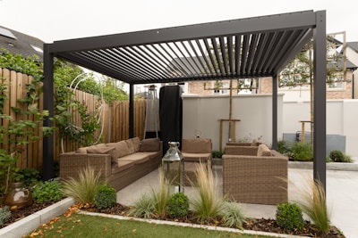 A modern outdoor seating area with a pergola structure made of metal beams. The space features wicker sofas with brown cushions arranged in an L-shape. Various plants and shrubs are placed around the seating area, and a wooden fence encloses the space. A large lantern sits on the ground, and the overall setting appears tidy and well-maintained.
