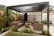 A modern outdoor seating area with a pergola structure made of metal beams. The space features wicker sofas with brown cushions arranged in an L-shape. Various plants and shrubs are placed around the seating area, and a wooden fence encloses the space. A large lantern sits on the ground, and the overall setting appears tidy and well-maintained.