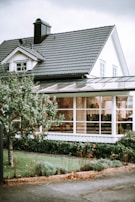 A picturesque suburban house with white wooden siding and a dark grey tiled roof. The structure features a quaint dormer window and a prominent chimney. In the foreground, a lush tree with visible red fruit stands next to a neatly trimmed lawn and bushes. Large windows and a possibly enclosed patio or sunroom are visible, hinting at cozy interior decor.