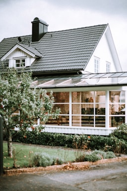 A picturesque suburban house with white wooden siding and a dark grey tiled roof. The structure features a quaint dormer window and a prominent chimney. In the foreground, a lush tree with visible red fruit stands next to a neatly trimmed lawn and bushes. Large windows and a possibly enclosed patio or sunroom are visible, hinting at cozy interior decor.