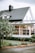 A picturesque suburban house with white wooden siding and a dark grey tiled roof. The structure features a quaint dormer window and a prominent chimney. In the foreground, a lush tree with visible red fruit stands next to a neatly trimmed lawn and bushes. Large windows and a possibly enclosed patio or sunroom are visible, hinting at cozy interior decor.
