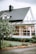A picturesque suburban house with white wooden siding and a dark grey tiled roof. The structure features a quaint dormer window and a prominent chimney. In the foreground, a lush tree with visible red fruit stands next to a neatly trimmed lawn and bushes. Large windows and a possibly enclosed patio or sunroom are visible, hinting at cozy interior decor.