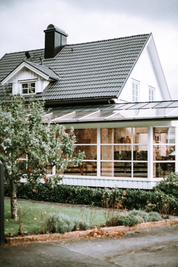 A picturesque suburban house with white wooden siding and a dark grey tiled roof. The structure features a quaint dormer window and a prominent chimney. In the foreground, a lush tree with visible red fruit stands next to a neatly trimmed lawn and bushes. Large windows and a possibly enclosed patio or sunroom are visible, hinting at cozy interior decor.