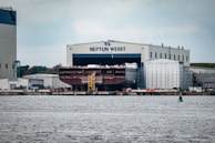 A large shipyard facility with a massive ship under construction inside a building marked 'Neptun Werft'. The ship, only partially visible, is located beside industrial structures and surrounded by scaffolding. The scene includes a vibrant waterway in the foreground and several smaller industrial buildings around the yard.