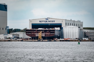 A large shipyard facility with a massive ship under construction inside a building marked 'Neptun Werft'. The ship, only partially visible, is located beside industrial structures and surrounded by scaffolding. The scene includes a vibrant waterway in the foreground and several smaller industrial buildings around the yard.