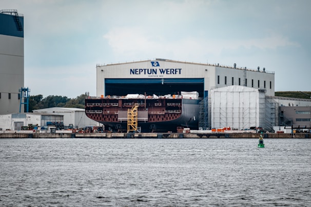 A large shipyard facility with a massive ship under construction inside a building marked 'Neptun Werft'. The ship, only partially visible, is located beside industrial structures and surrounded by scaffolding. The scene includes a vibrant waterway in the foreground and several smaller industrial buildings around the yard.