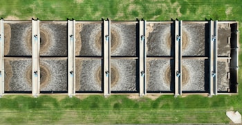 An aerial view of a wastewater treatment facility featuring eight large, rectangular sedimentation tanks arranged in two rows. Each tank contains circulating water, likely part of the treatment process. The tanks are surrounded by green grass, giving a structured and organized appearance.